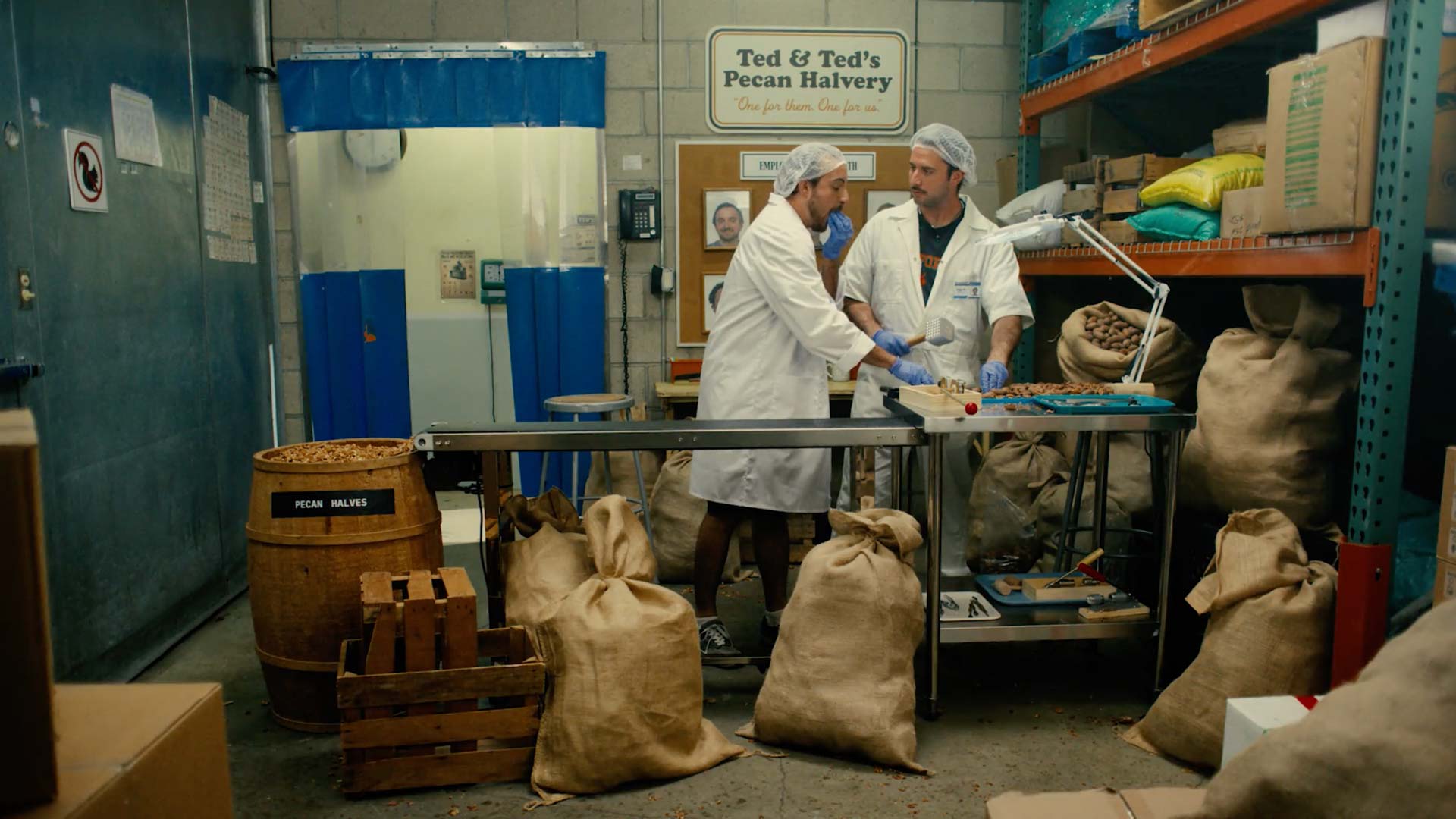 Still frame from "Other Half" tv spot of 2 men wearing white coats, gloves and hair nets standing at a conveyer belt emptying pecans into a barrel in a crowded back room with bags of pecans and a sign saying "Ted & Ted's Pecan Halvery"